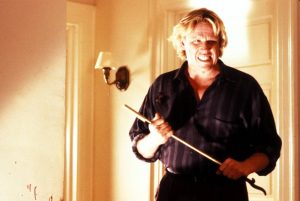A man with blonde hair and a striped shirt stands indoors gripping a gold cane, showing an intense facial expression. There is blood spatter visible on the wall to his left.