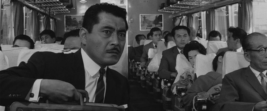 A man in a suit sits tensely on a crowded train, clutching a briefcase, while other passengers around him read newspapers or look forward.