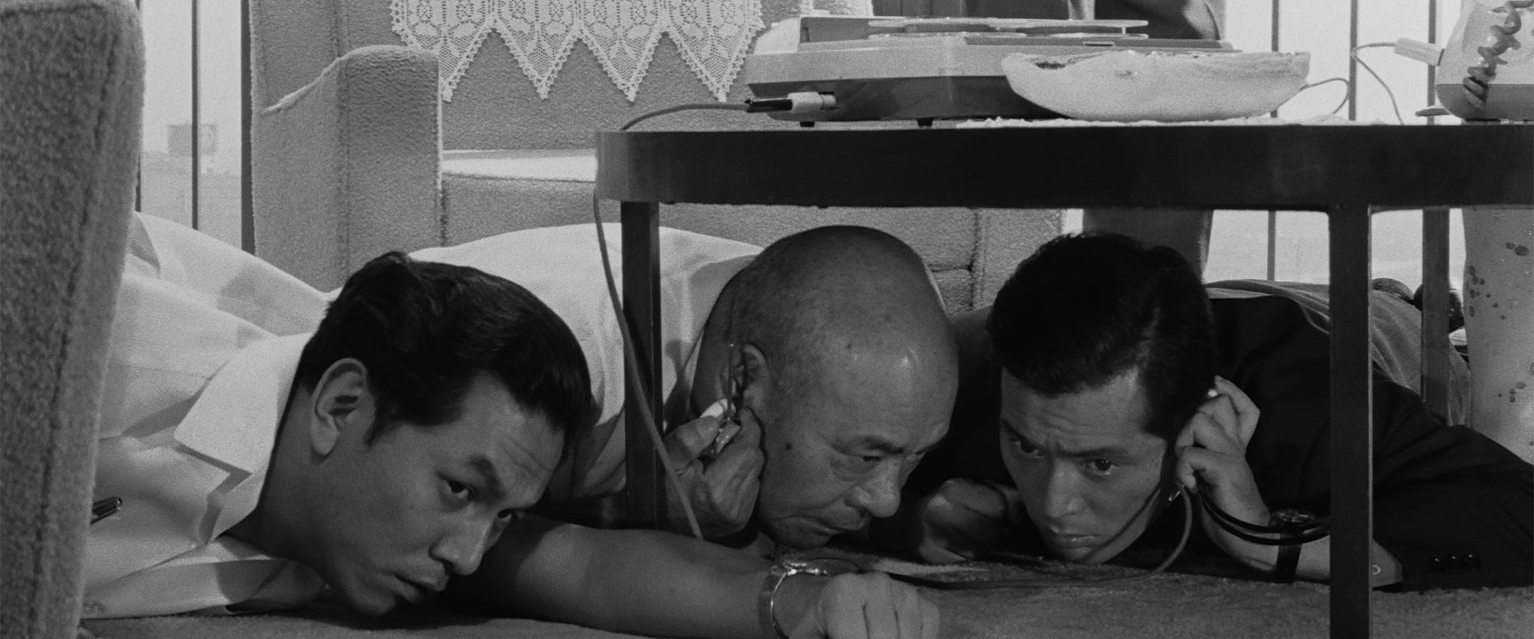 Three men lie on the floor under a table, each holding a stethoscope to the surface above them, appearing to listen intently.