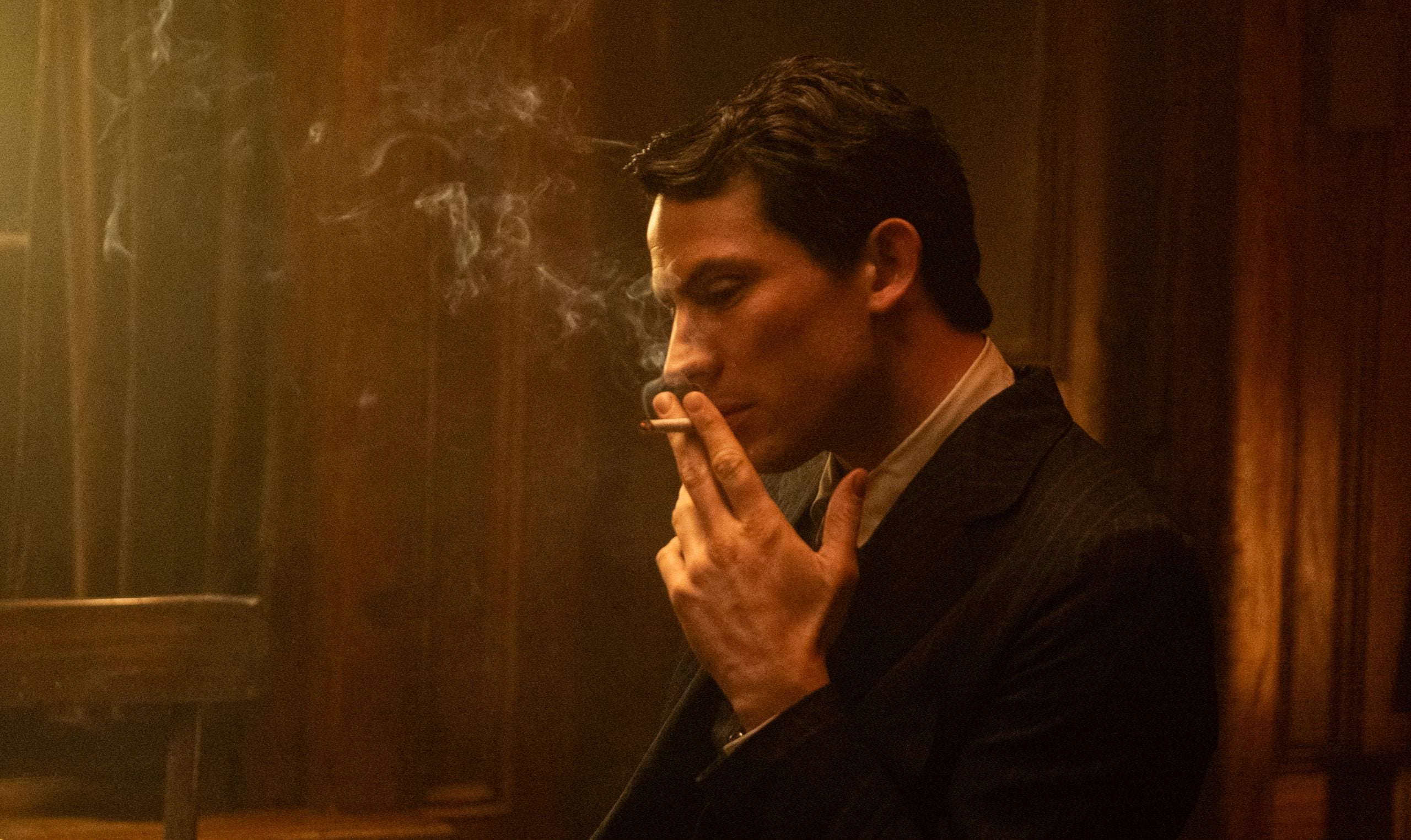 A man in a suit sits in a dimly lit room, holding a cigarette with smoke rising, looking contemplative.