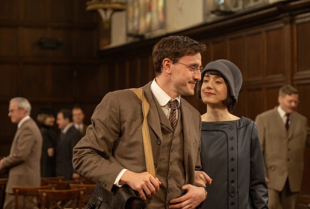 A man in a brown suit and glasses stands next to a woman in a blue dress and hat inside a wood-paneled room, with other people conversing in the background.