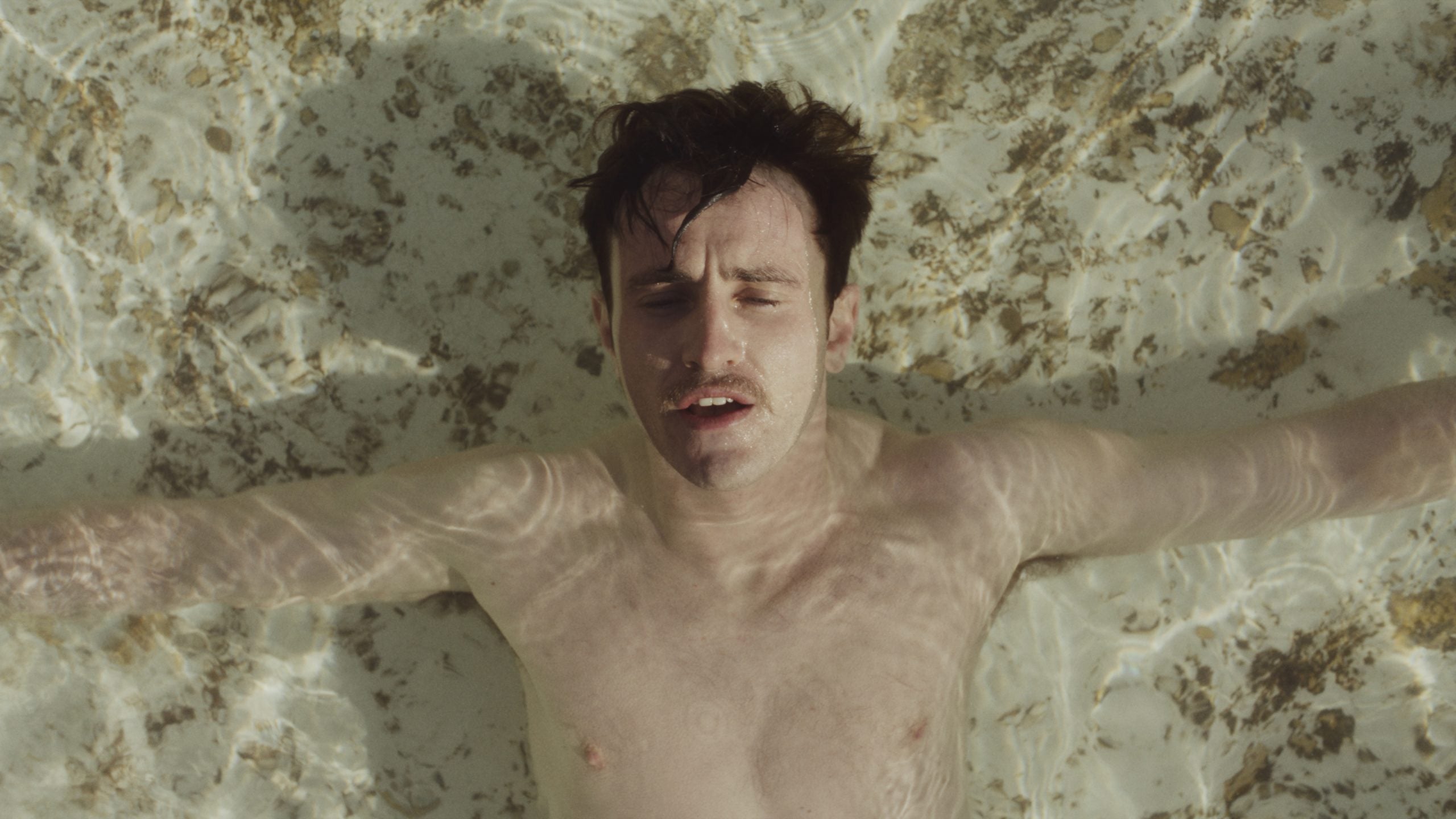 A man with short dark hair floats on his back in shallow, clear water with his arms outstretched, looking upward.