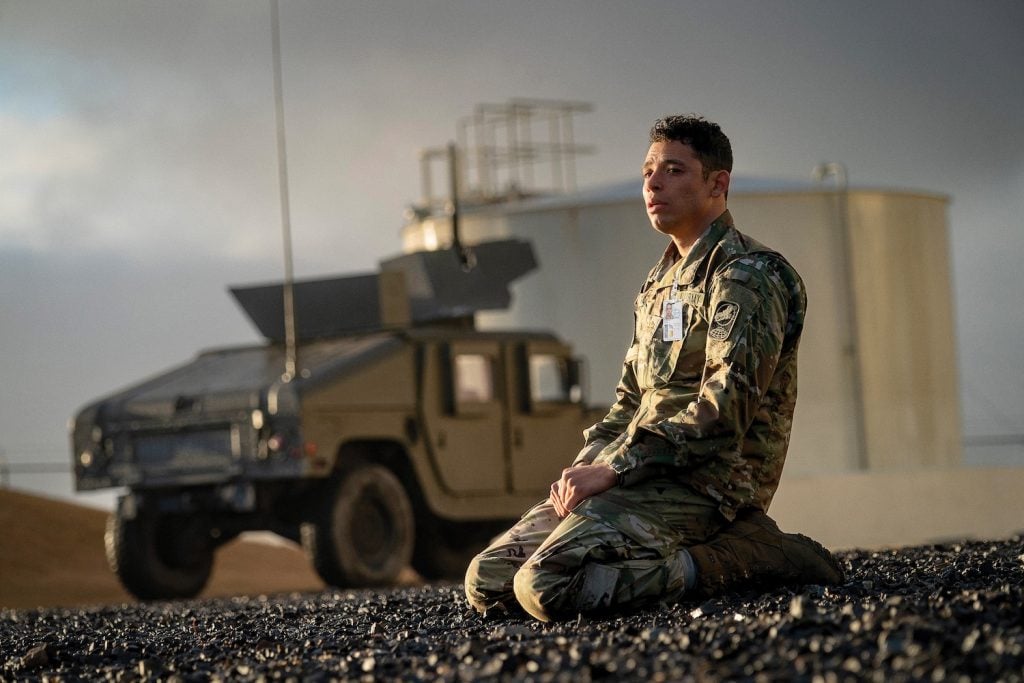 A person in military uniform kneels on rocky ground with a military vehicle and an industrial tank in the background.