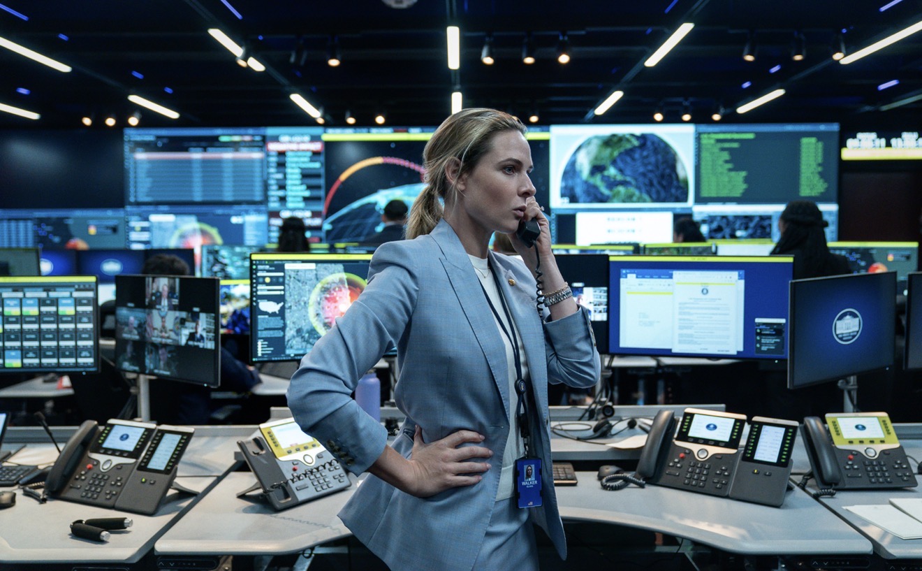 A woman in a suit stands in a control center, talking on the phone, surrounded by desks with computers, monitors, and telephones displaying data and maps.