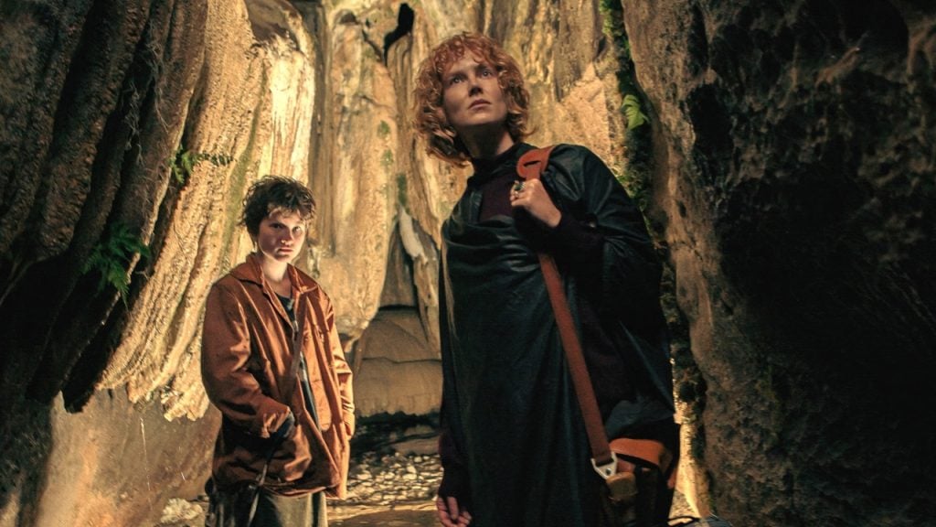 Two people stand inside a rocky cave, looking up toward the light. Both wear casual clothes and carry bags. The cave walls are textured and lit by natural light.