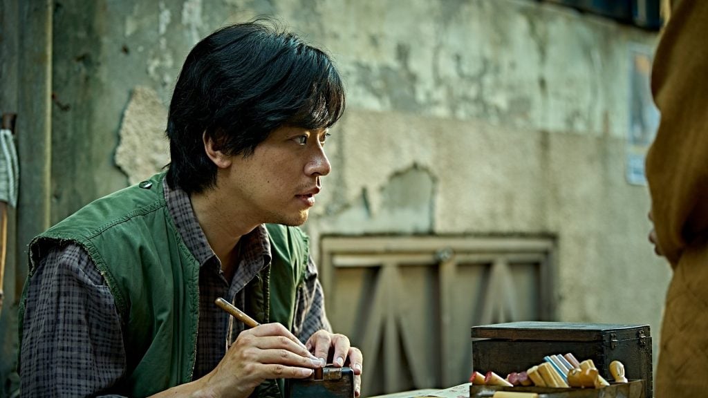 A man sits at a table, carving or working with tools, focused on his task. The background shows a weathered wall and a closed wooden door.