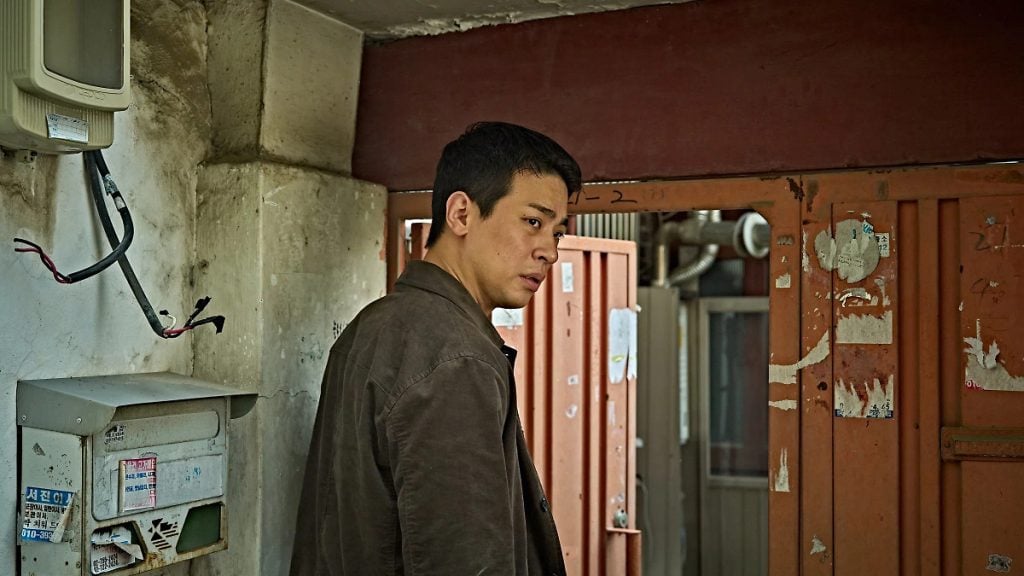 A man in a brown jacket stands in a narrow, weathered entryway with peeling paint and exposed wires, looking to his right.