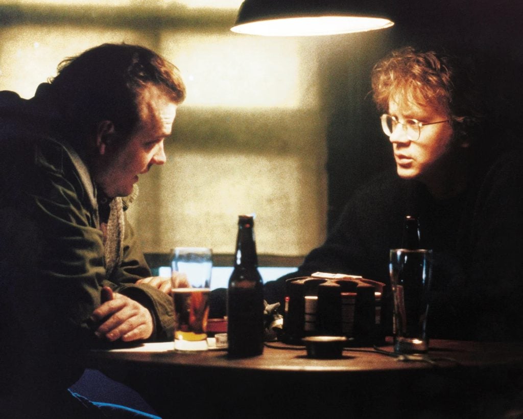 Two people sit at a round table in a dimly lit room, talking, with drinks and bottles in front of them under a hanging lamp.