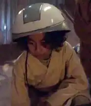 A young child wearing a large white helmet and beige clothing is crouched down indoors.