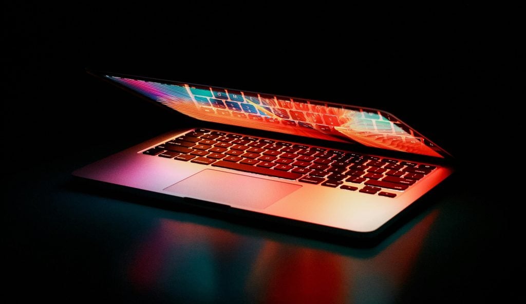 A partially closed laptop glows with colorful reflections from its screen and keyboard in a dark environment.