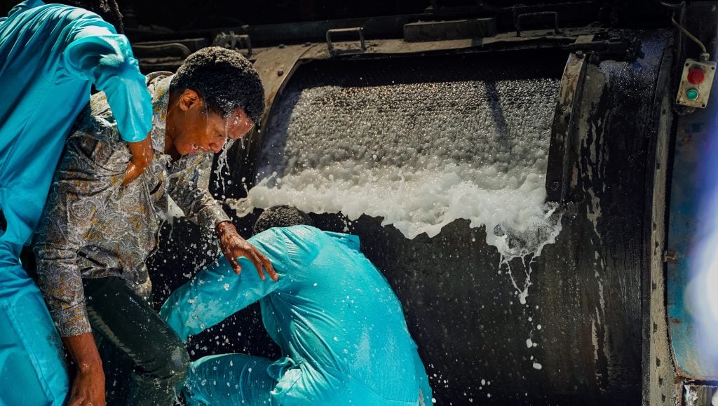 Three people, two in blue uniforms, are splashed by water pouring from a large industrial machine.