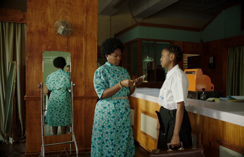 A woman in a blue patterned dress counts money while speaking to a girl in a school uniform holding a briefcase at a counter in a vintage-style room.
