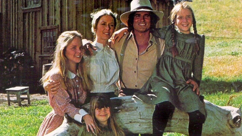 A family of five poses outdoors by a wooden log, with a barn in the background. The adults and children wear old-fashioned, prairie-style clothing.