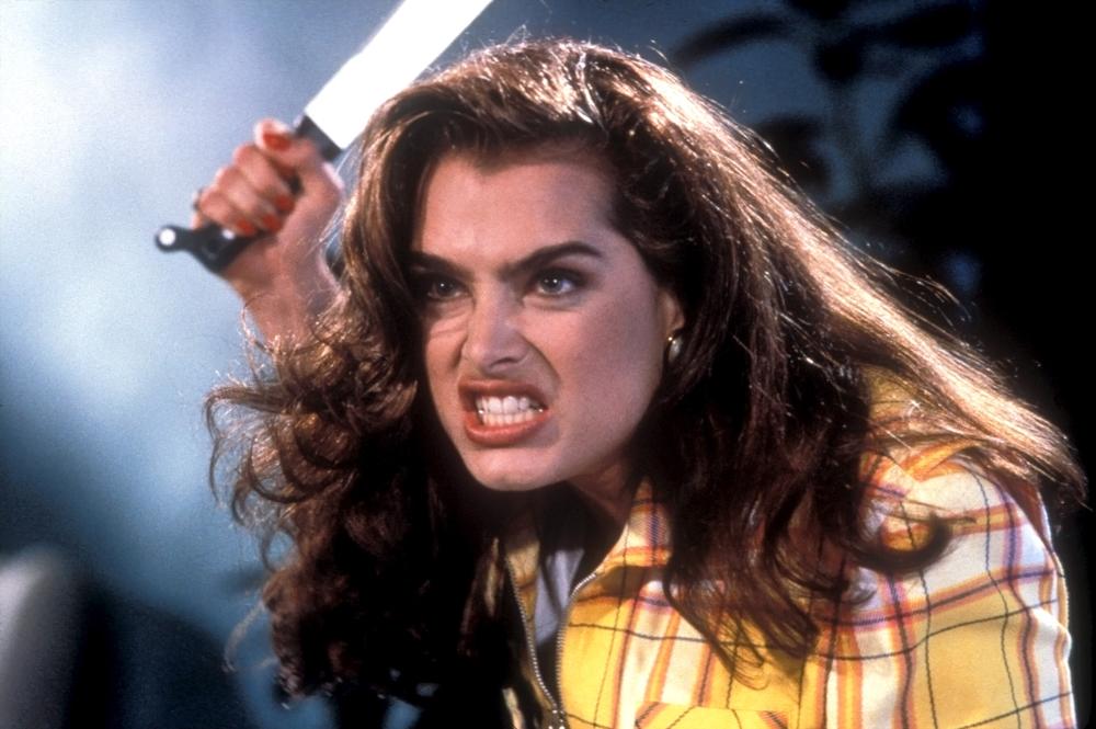 A woman with long brown hair and a yellow plaid shirt holds a large knife above her head, looking angry and intense.
