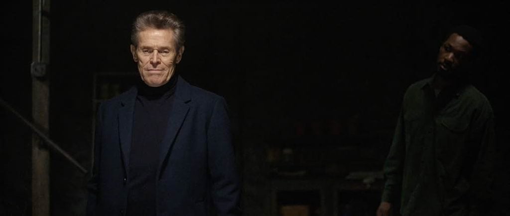 A man in a dark turtleneck and blazer stands in a dimly lit room, with another man partially visible in the background.