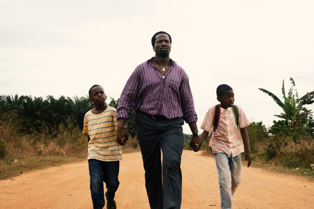 An adult man walks down a dirt road holding hands with two boys, one on each side, against a rural backdrop with trees and plants.