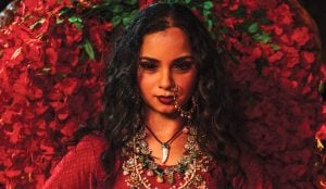 Woman with long curly hair wearing ornate jewelry and a red outfit stands in front of a backdrop of red flowers, looking directly at the camera.