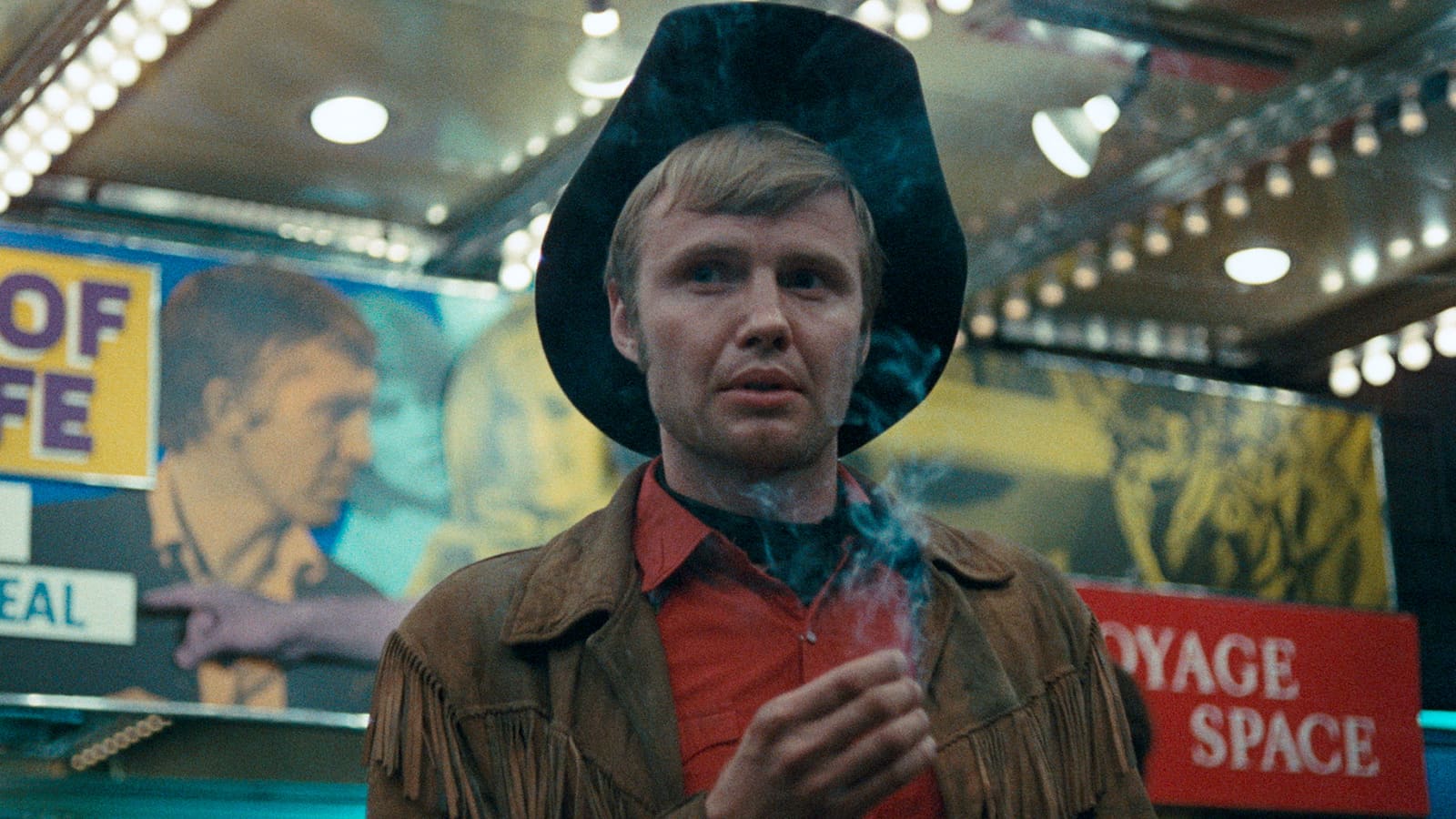 A man in a cowboy hat and fringe jacket stands under bright lights, holding a cigarette, with colorful signs and a reflection of his face in the background.