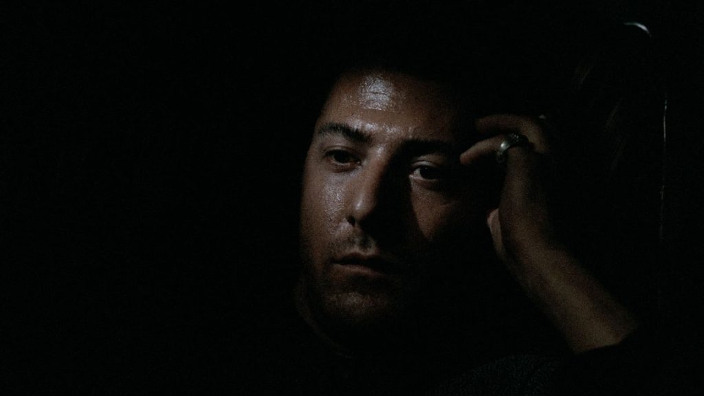 A man sits in darkness with a serious expression, resting his head on his hand. Only part of his face and hand are illuminated by dim light.