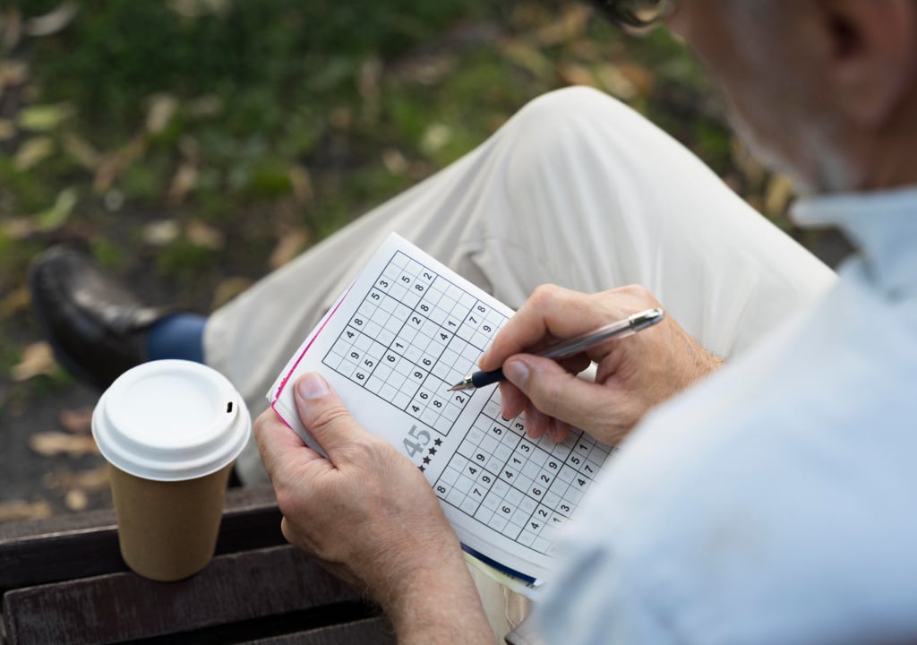 A person sitting on a bench completes a sudoku puzzle in a notebook with a pen, with a disposable coffee cup placed nearby.