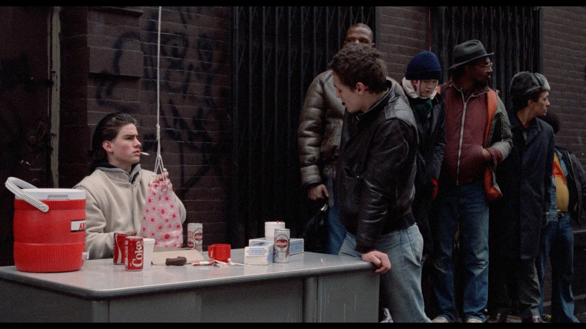 A person sits at a desk with drinks and snacks, wearing a light hoodie, while another person in a leather jacket leans on the desk. Several people stand in line against a brick wall.