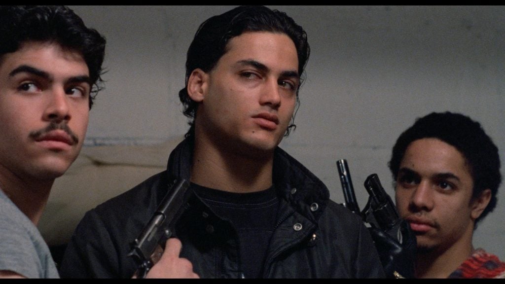 Three young men stand close together indoors; the two on the sides are holding handguns, and all have serious expressions.