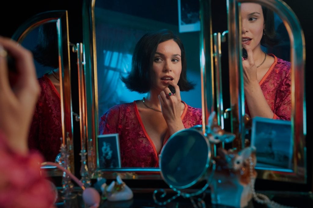A woman in a pink dress applies lipstick while looking at herself in a tri-fold vanity mirror, surrounded by jewelry and personal items.