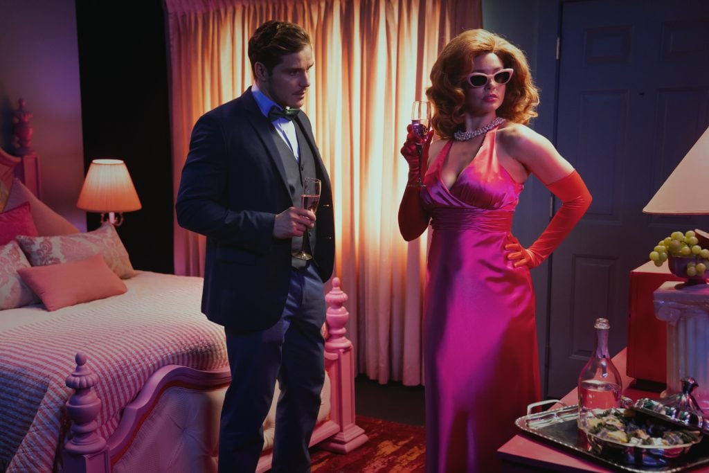 A man in a suit and a woman in a pink evening gown with sunglasses and gloves stand in a hotel room, holding drinks, with a bed and table of food in the background.