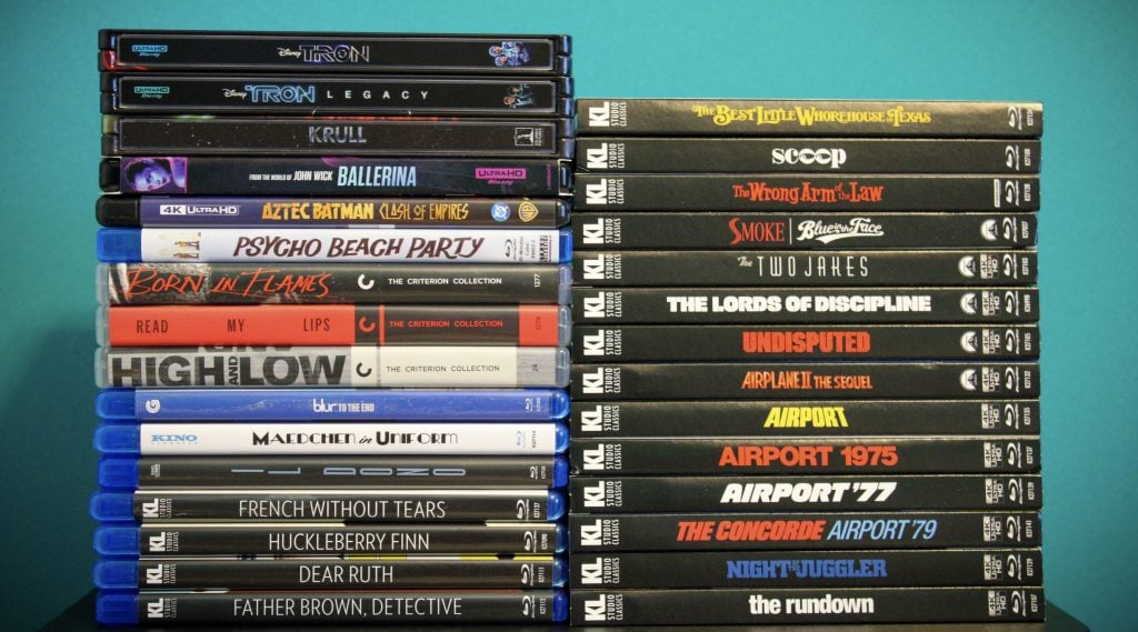 A stack of assorted DVD and Blu-ray movie cases is arranged against a teal background, with titles visible on the case spines.