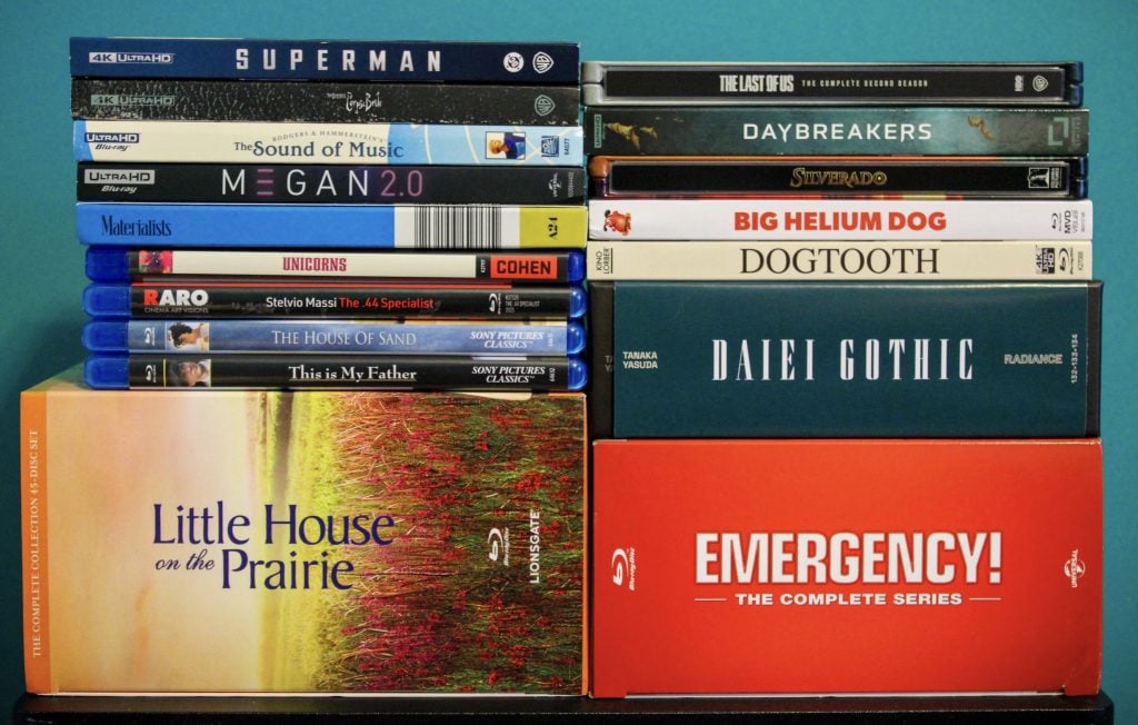A collection of DVD and Blu-ray cases, including titles like Superman, Little House on the Prairie, Dogtooth, and Emergency! The Complete Series, stacked against a blue background.