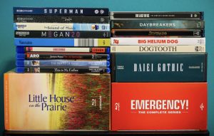 A collection of DVD and Blu-ray cases, including titles like Superman, Little House on the Prairie, Dogtooth, and Emergency! The Complete Series, stacked against a blue background.