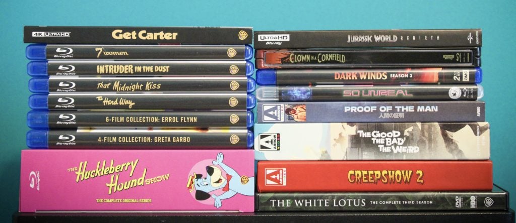 A stack of Blu-ray and DVD cases, including "Get Carter," "Jurassic World: Dominion," "The Huckleberry Hound Show," and several others, arranged against a teal background.