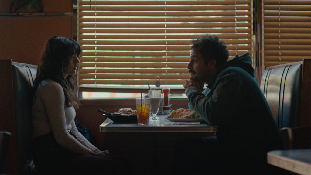 A woman and a man sit across from each other in a diner booth, having a conversation over food and drinks with window blinds in the background.