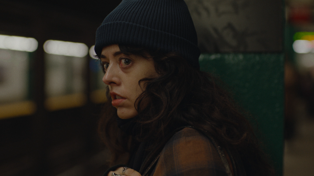 A woman in a dark beanie and plaid shirt stands next to a green pillar in a subway station, looking over her shoulder with a serious expression.
