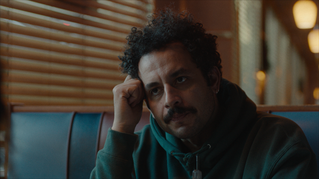 A man with curly hair and a mustache sits in a booth at a diner, resting his head on his hand, looking slightly pensive.