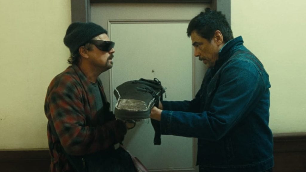 Two men stand in a hallway facing each other, holding opposite ends of a worn boot, with a closed door behind them.