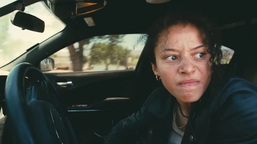A person with a tense expression sits in the driver's seat of a car, looking out the open door.