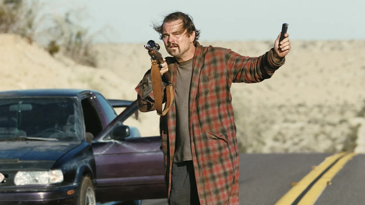 A man in a plaid robe stands on a deserted road holding a rifle and a handgun, with a car parked behind him and desert landscape in the background.