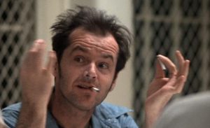 Man with messy hair and cigarette in mouth gestures with both hands while sitting indoors, wearing a light blue shirt.