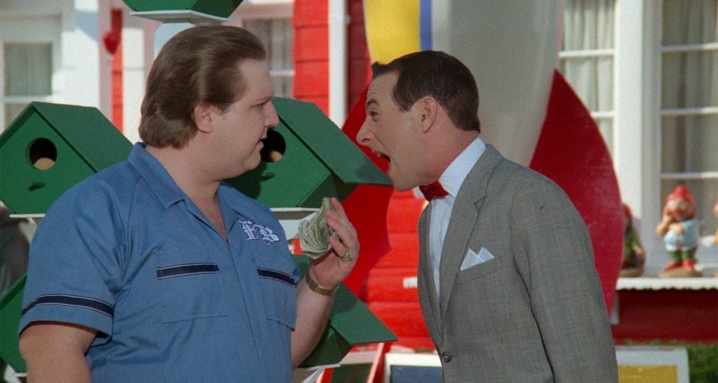 Two men stand outside; one in a blue shirt holds money while the other in a gray suit and red bowtie appears to argue or shout at him. Green birdhouses and colorful objects are in the background.