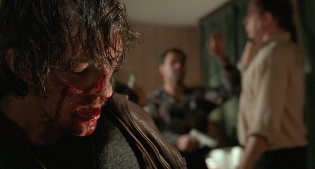 A man with a bloodied face looks downward in the foreground, while two other men stand in the blurred background, one with his arm raised.