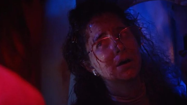 A woman with blood on her face and glasses looks upward with a distressed expression under red and blue lighting.