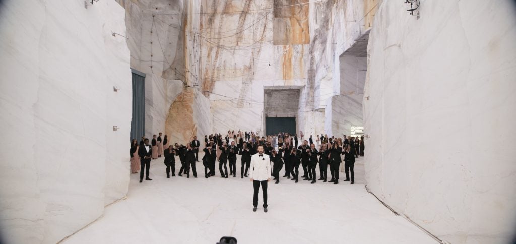 A man in a white jacket stands in the foreground of a large, white, marble-walled hall with a crowd of formally dressed people gathered behind him.