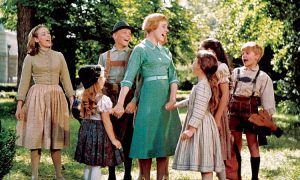 A woman and seven children stand outdoors in a group, holding hands and singing together, surrounded by trees and greenery.