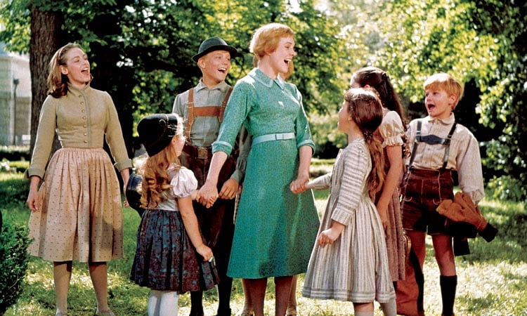 A woman and seven children stand outdoors in a group, holding hands and singing together, surrounded by trees and greenery.