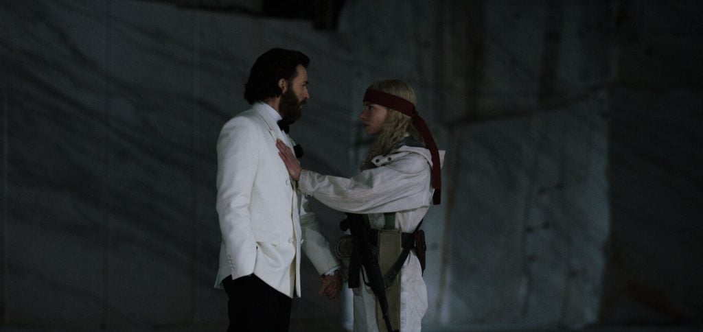 A man in a white suit stands facing a woman in tactical clothing and a red headband, who is touching his chest in a dimly lit, marble-walled setting.