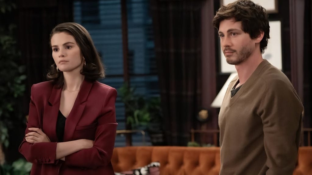 A woman in a maroon blazer stands with her arms crossed next to a man in a brown sweater in a warmly lit living room.