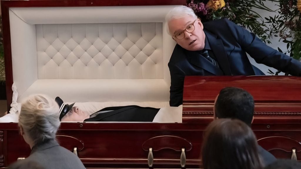 A man leans over an open casket at a funeral, addressing seated attendees while the deceased lies inside dressed in black.