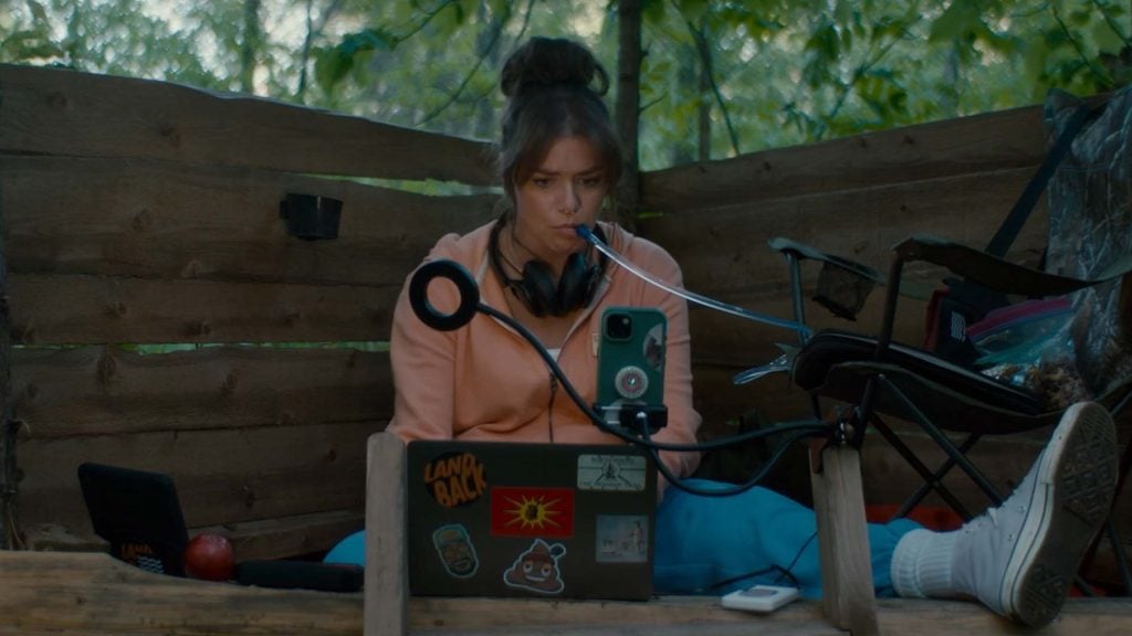 A person sits against a wooden wall outdoors, using a laptop with stickers, a phone mounted on a stand, and a headset around their neck.