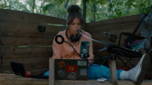 A person sits against a wooden wall outdoors, using a laptop with stickers, a phone mounted on a stand, and a headset around their neck.